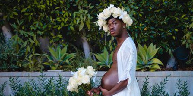 Lil Nas X in bridal wear, with a white robe, a white flower crown and bouquet, and a pregnant belly.