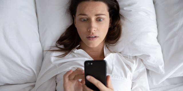 A person with long brown hair in a white shirt is in a bed with white sheets. They hold a black phone and look concerned.