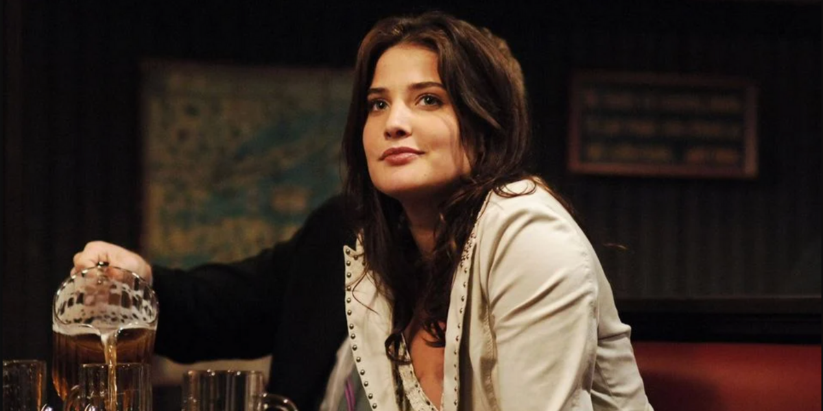 Image shows Robin, sitting in a bar wearing a cream colored blazer looking off into the distance with a slight smirk