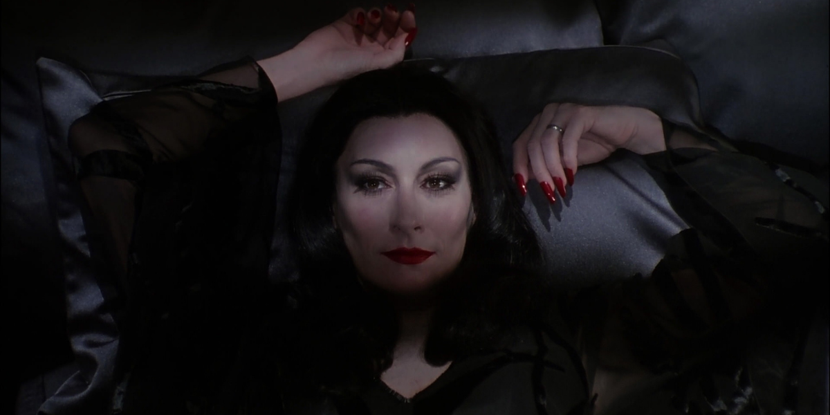 Image shows Morticia Addams laying in bed with her arms draped around her, pointed red nails and long flowing black hair staring deeply into the camera.