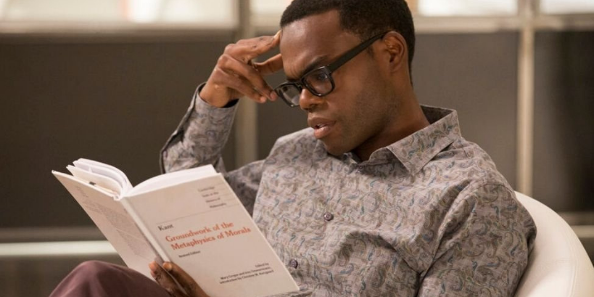 Image shows Chidi, A Black man with a short fade, reading a book. He is wearing Black thick glasses and a slight patterned gray button down