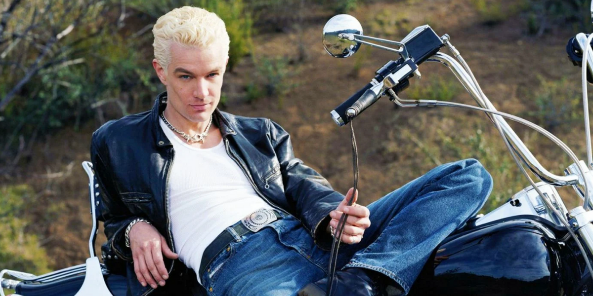 Image shows a blonde haired Spike leaning back on a motorcycle wearing a leather jacket and a white tank top