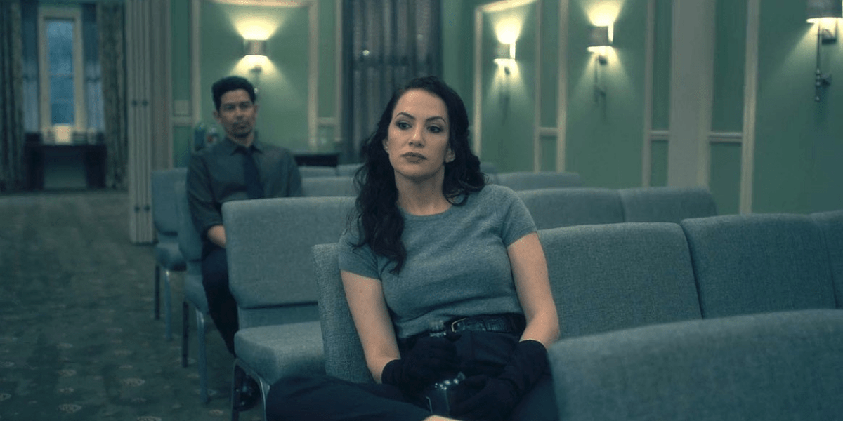 Image shows Theo sitting on the last seat in a row of chairs wearing a deep grey tshirt, black pants and gloves while giving a tilted side eye glance to someone