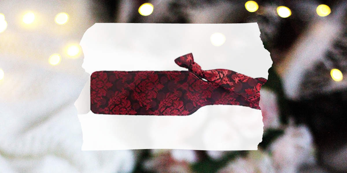 Sex toy gift guide: Image shows a rectangle paddle with deep purple and red paisley style print overlaid on a photo of roses and Christmas lights.