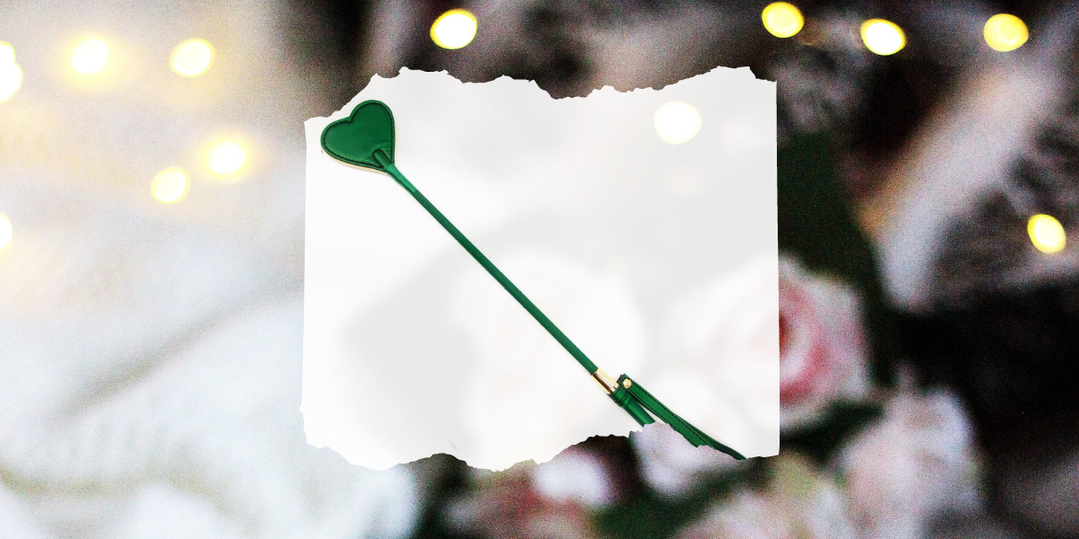 Sex toy gift guide: Image shows A emerald green riding crop with a heart shaped top and gold accents overlaid on a photo of roses and Christmas lights.