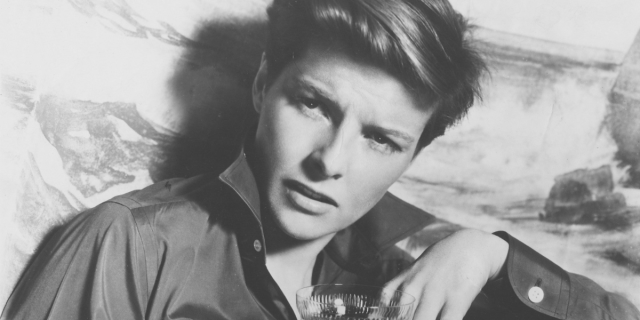 Image shows Katherine Hepburn with a perfectly cut short haircut, wearing a buttondown shirt slightly open and looking off to the side with a sexy squint