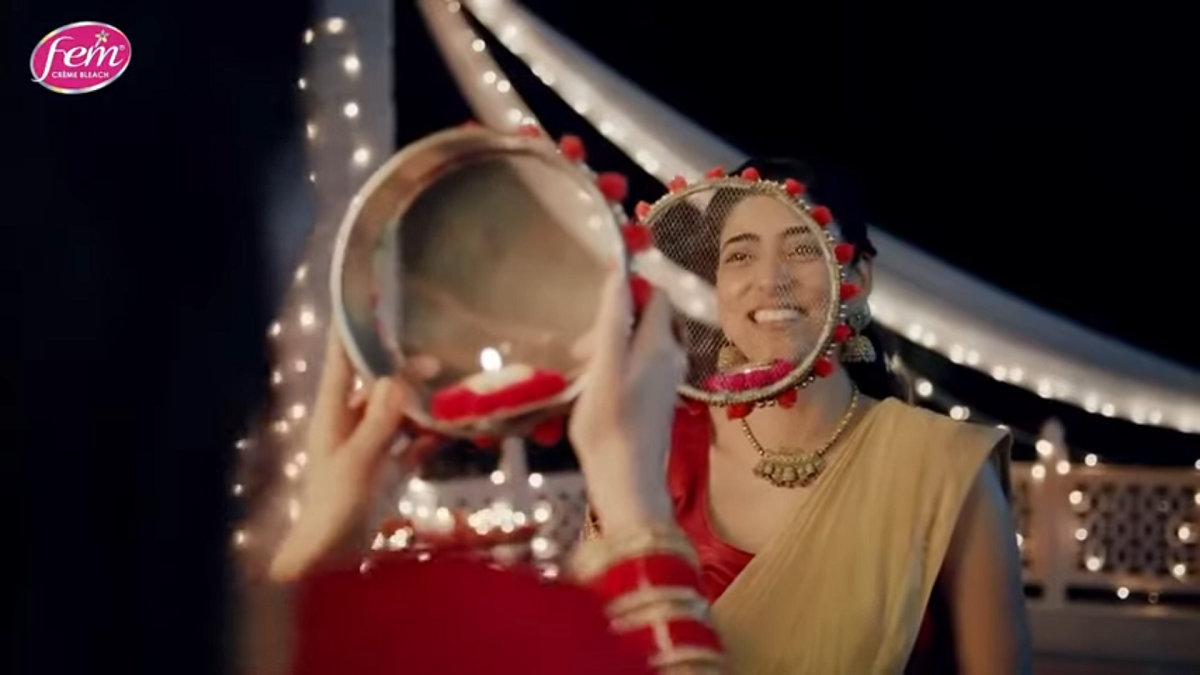 A mostly off screen woman wearing red bangles holds a decorated sieve with a candle inside. Looking through the sieve is another woman holding a sieve, smiling brightly and looking back at the off screen woman.