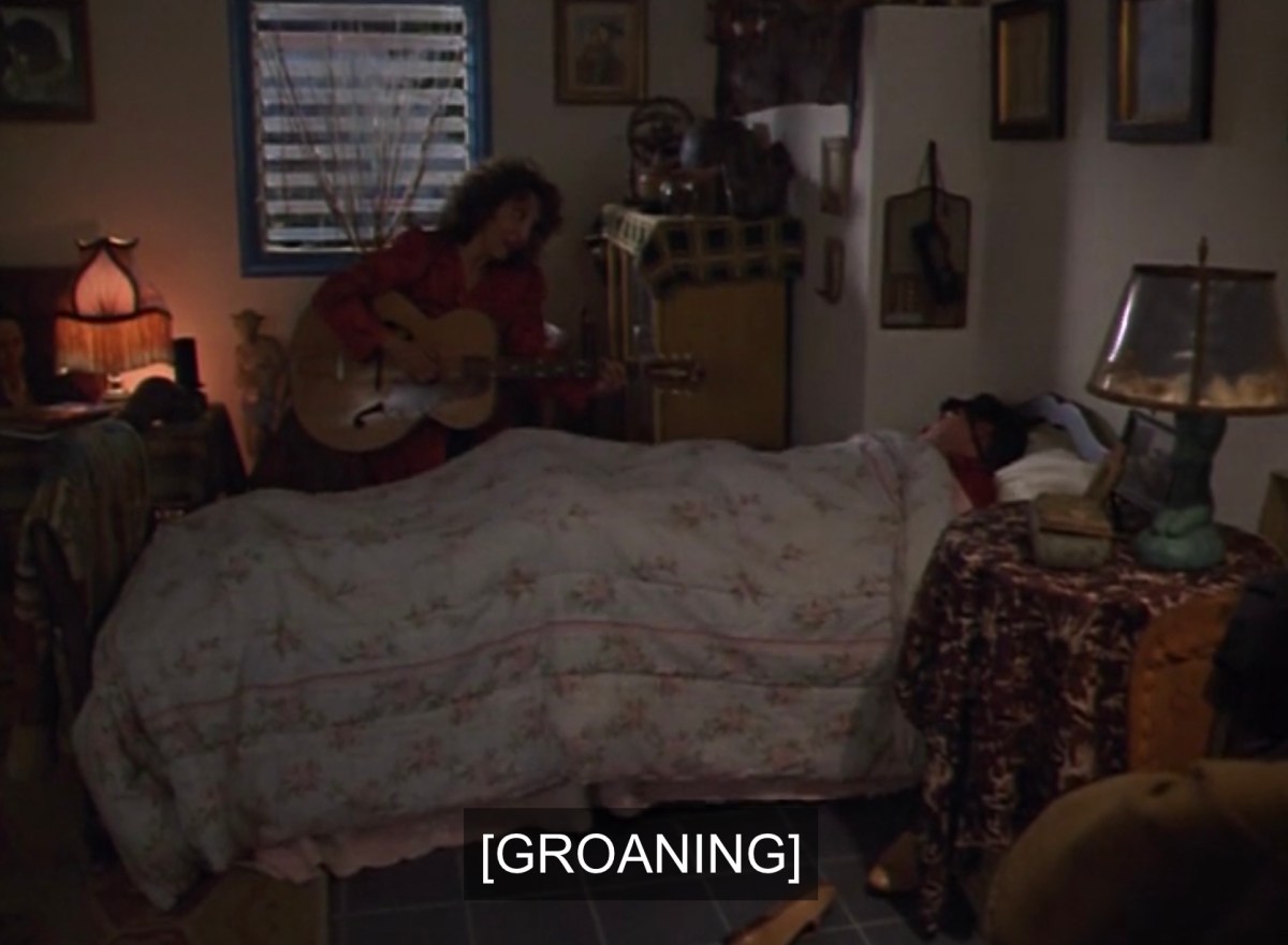 Reed in bed while Bitzy plays guitar, caption reads [GROANING]