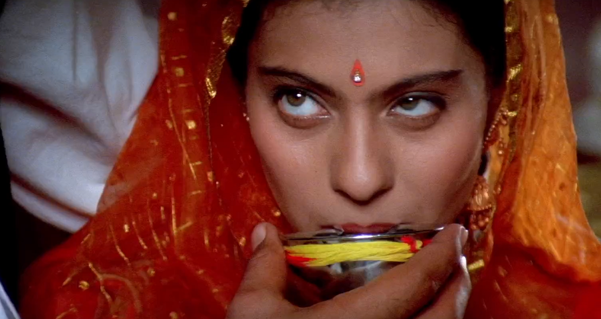 Simran sips from a silver bowl decorated yellow string that is being held up to her. She is looking up with a knowing glance.