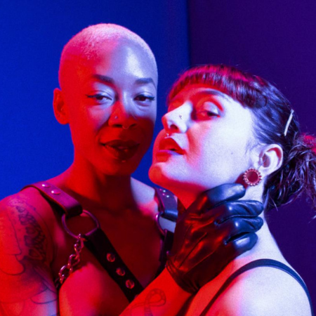 A shirtless person with buzzed, bleached blonde hair wears a chest harness and leather gloves. They wrap their hand around the throat of another person, who has short bangs and long brown hair that is pinned up.