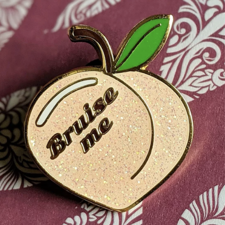 A glittery enamel pin shaped like a peach reads, "Bruise me." It is against a red background with a white swirling pattern.