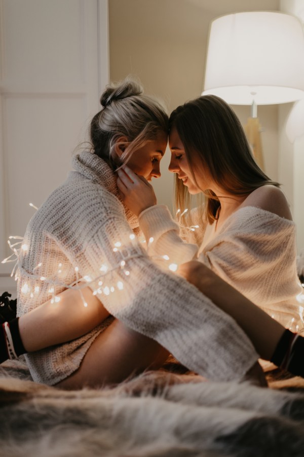two white queers cuddle in bed, wrapped in string lights