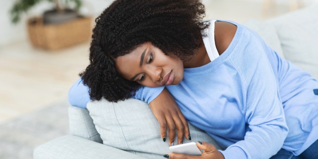 A person with dark brown skin and black curly hair wears a light blue shirt with a white tank underneath. They lean on a couch and look at their phone.