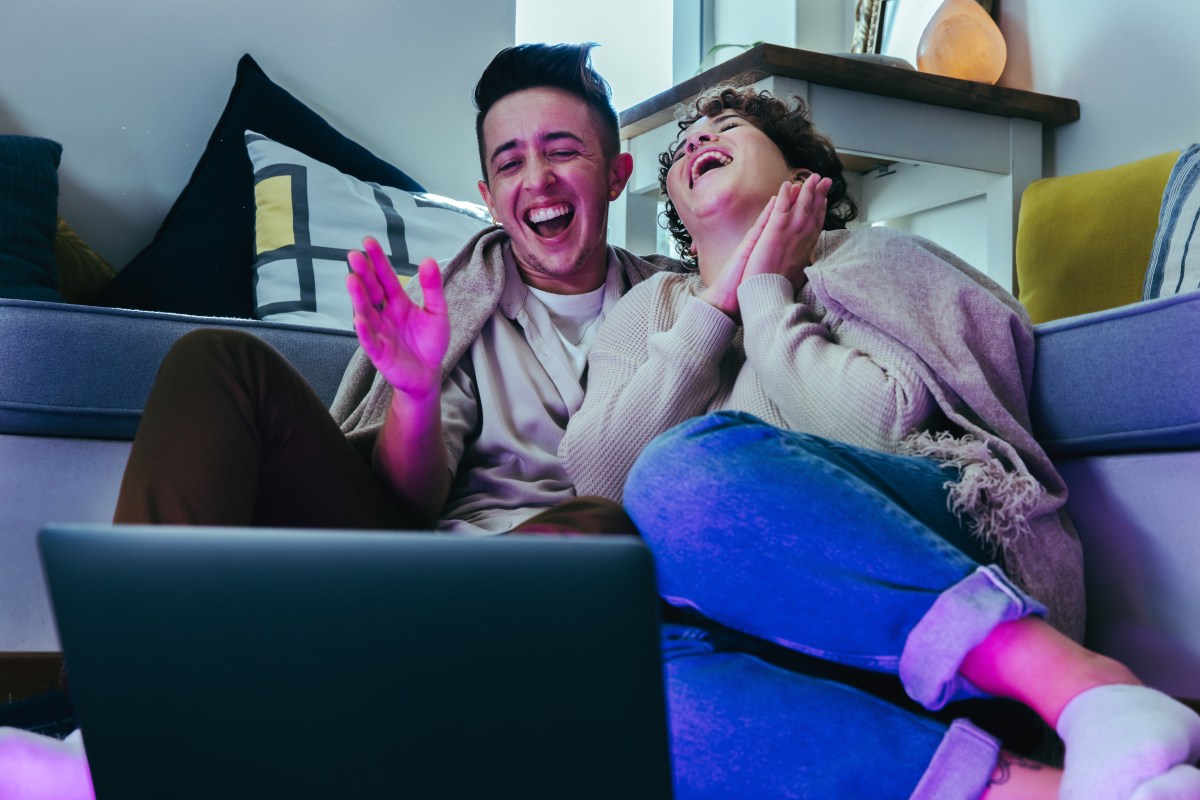 two queers laugh together on a couch while watching a laptop screen