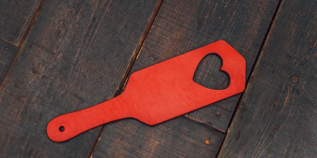 A red leather paddle with a heart-shaped cutout against a wood-paneled background