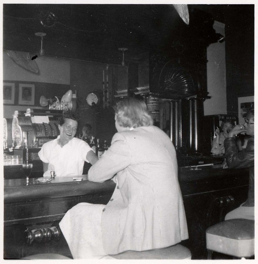 a black and white photo of the interior of a bar, where the smiling white-shirted bartander Grace Miller serves a woman sat on a stool at the bar with her back to the camera. To the right of the shot we glimpse butch-looking women smoking at the bar.
