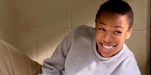 Image shows Poussey Washington, a Black masc lesbian, smiling into the camera while looking off to the right. She is propped up on her bed and wearing a grey sweatshirt and khaki pants.