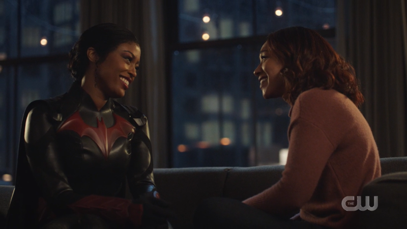 Ryan Wilder, dressed as Batwoman, and Iris West sitting on the couch smiling at each other.