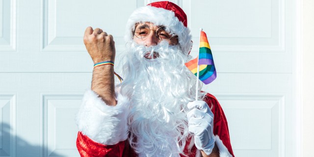A man in a santa suit holds up a rainbow flag in one hand and a balled fist of triumph with a rainbow bracelet in the other