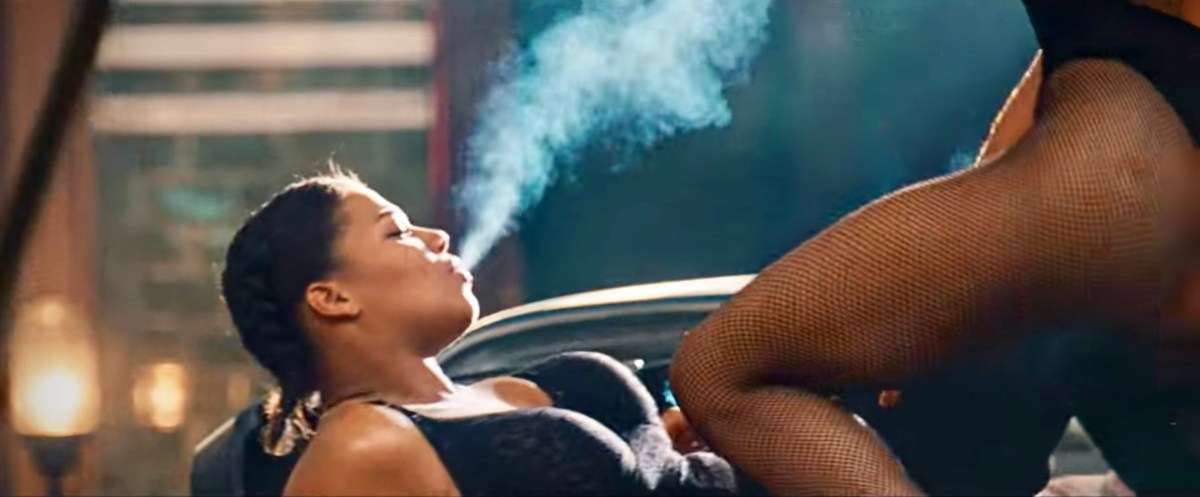 Image shows Cleo, a Black masc lesbian played by Queen Latifah, laying backwards on her car while blowing smoke out of her mouth. There are the legs of a femme person, covered in fishnet stockings, draped over her abdomen. Cleo is wearing lingerie.