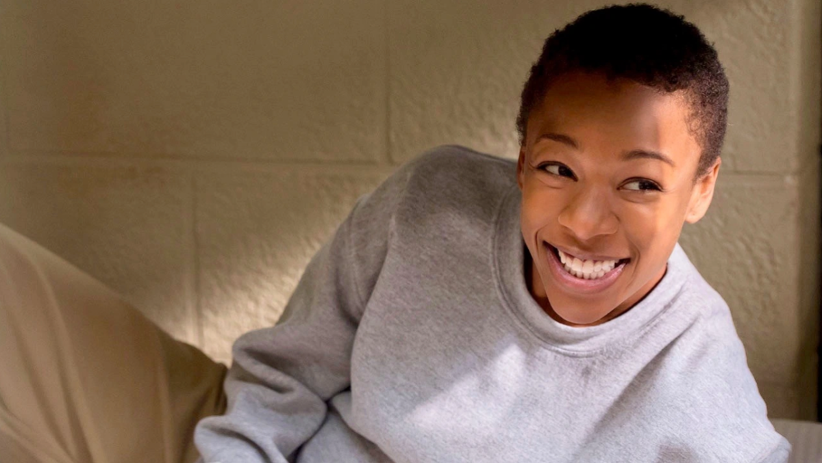 Image shows Poussey Washington, a Black masc lesbian, smiling into the camera while looking off to the right. She is propped up on her bed and wearing a grey sweatshirt and khaki pants.
