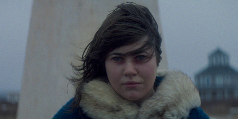 A close up on Teagan Johnston wearing red eye make up, their hair blowing in the wind, and a winter coat around them.