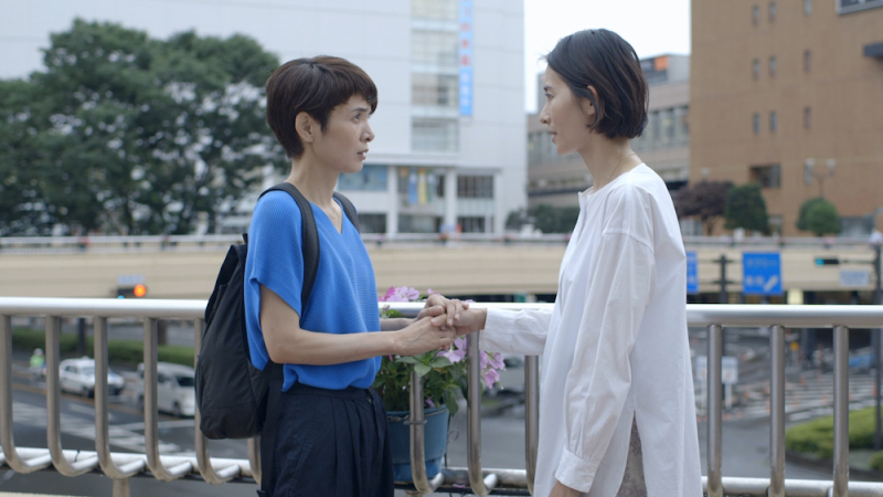 lesbian movies 2021 list still: Fusako Urabe and Aoba Kawai stand on an elevated train platform holding hands.