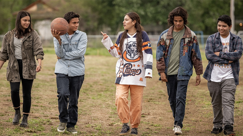 The cast of Reservation Dogs walking through a field holding a basketball and talking