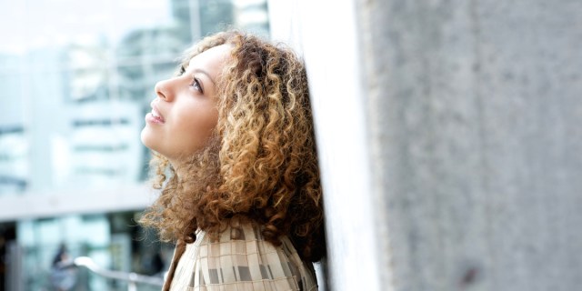 young woman with curly hair leans against a city wall and looks up at the sky, deep in thought
