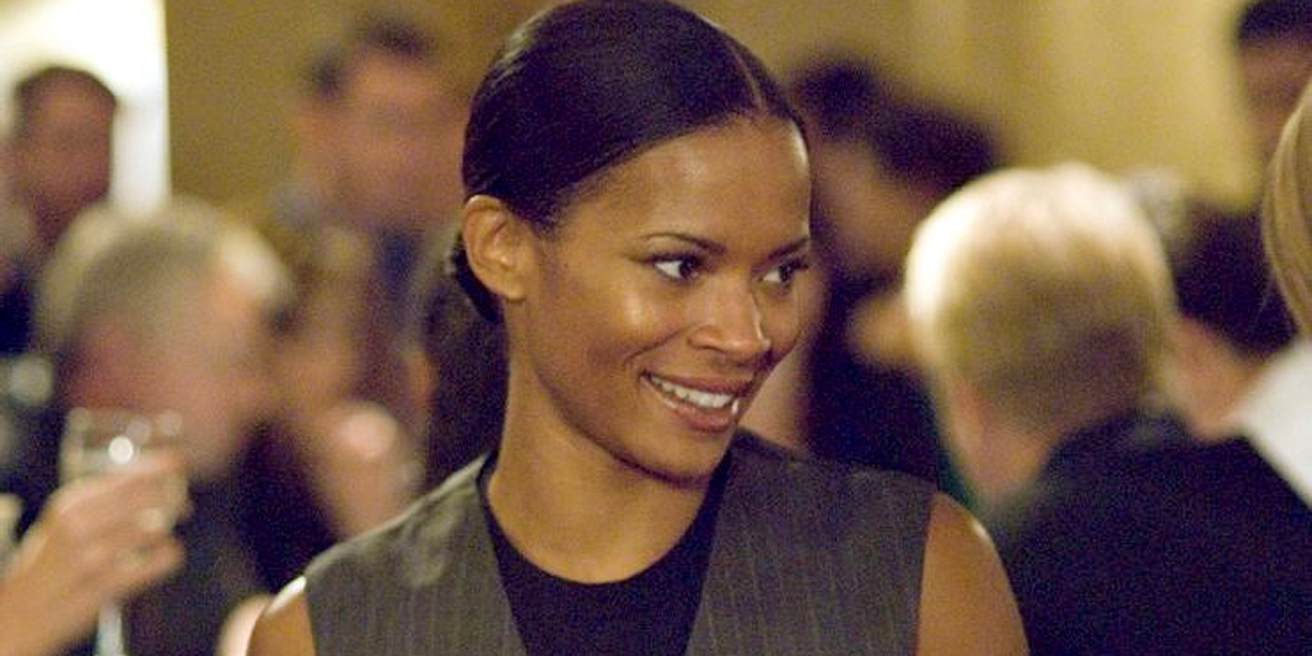 Image shows Tasha Williams, a Black lesbian with a low bun and severe hair part, from The L Word looking over her left shoulder and smiling to the camera. Tasha has on a pinstripe grey vest and black tank top. 