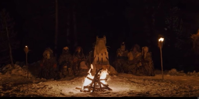 Still from Yellowjackets: A group of people gather in the dark behind a fire. There are two lit torches on either side of them. All of the people are wearing face coverings.