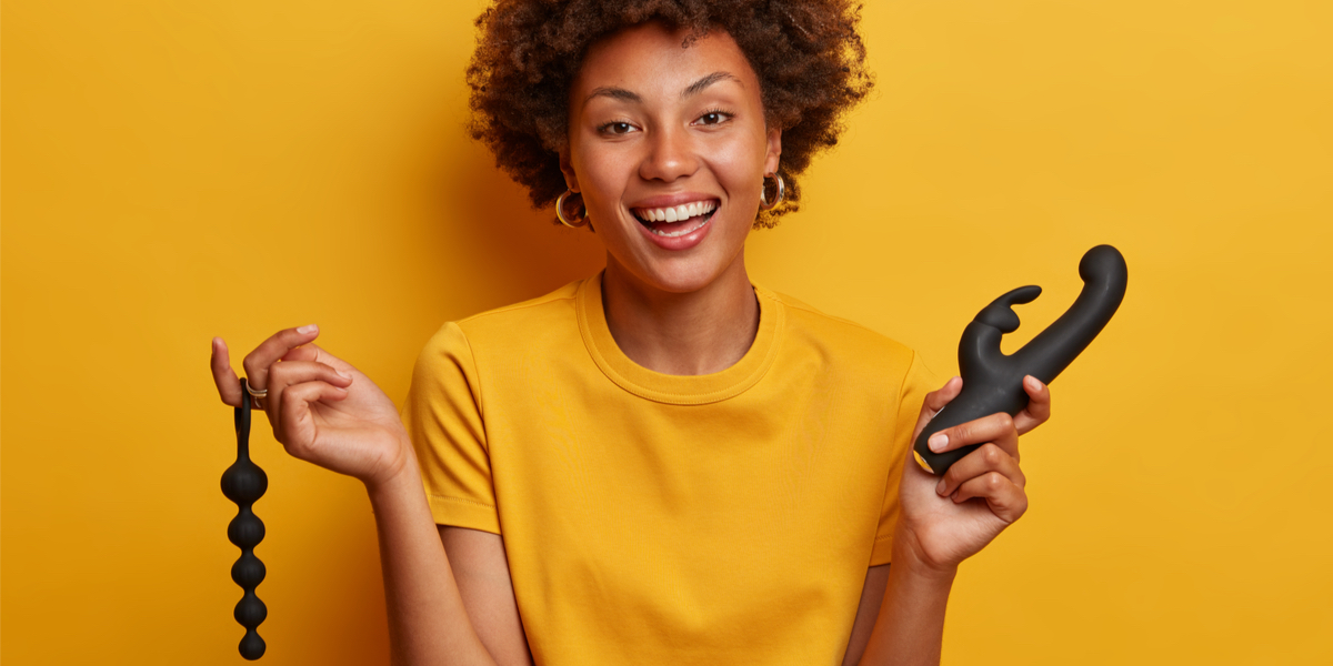 A black woman with an afro is wearing a yellow shirt and holding black sex toys