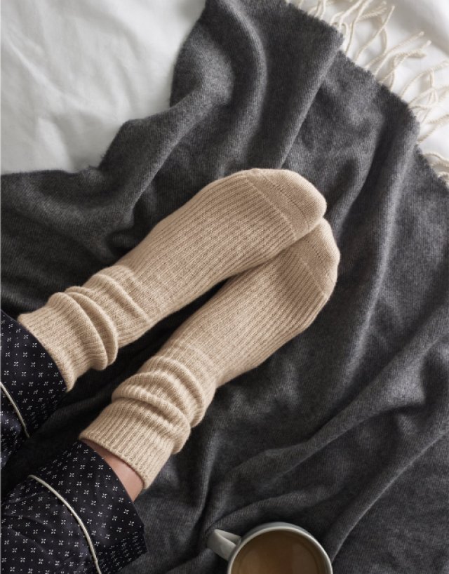 Bone colored socks against a soft dark grey blanket with a cup of tea in the corner