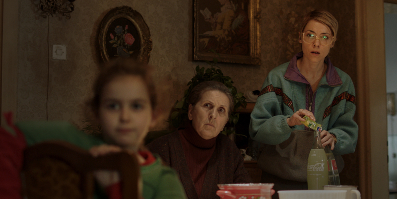 A young woman in big glasses stands pouring drink mix and looking in the same direction as the old woman next to her and he child in the foreground.