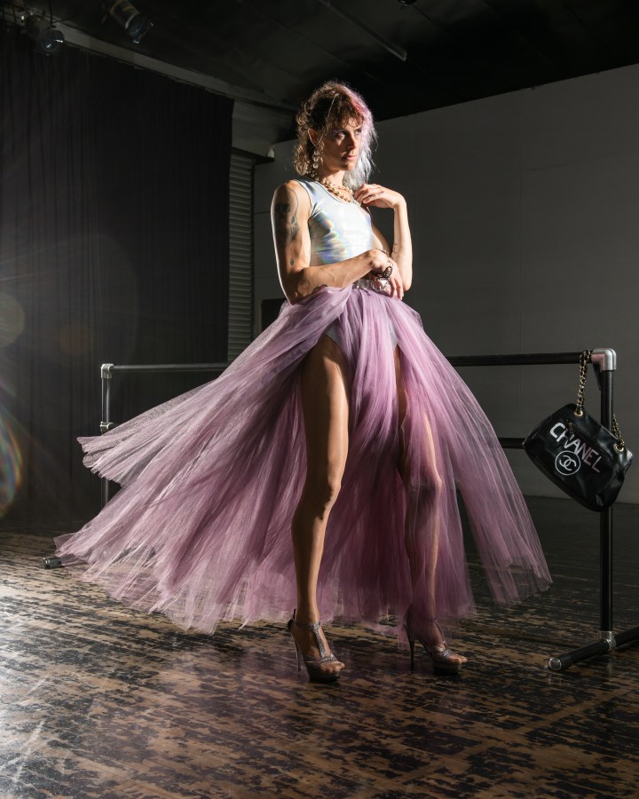 CJ Kitten Miller, the author of this essay, poses in a long purple tulle skirt and a shiny tank top. Her hair is pink and her heels are high. Three in a series of five.
