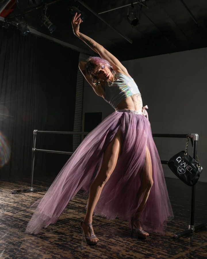 CJ Kitten Miller, the author of this essay, poses in a long purple tulle skirt and a shiny tank top. Her hair is pink and her heels are high. Five in a series of five.