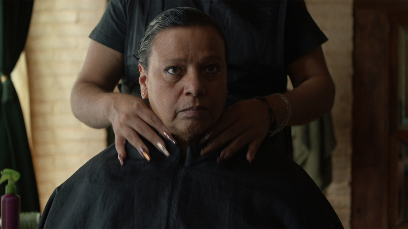 A butch woman with straight hair stairs forward as another woman with long nails prepares to cut her hair.