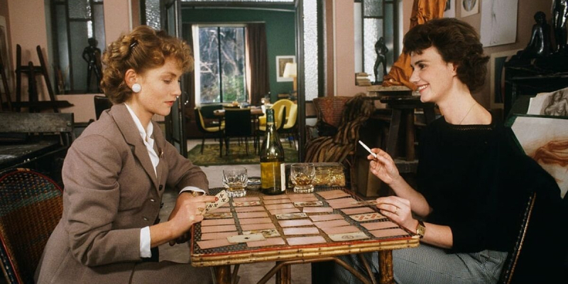 Miou-Miou and Isabelle Huppert sit across from a table playing cars. Huppert is looking down holding a card. Miou-Miou is holding a cigarette and looking at Huppert.