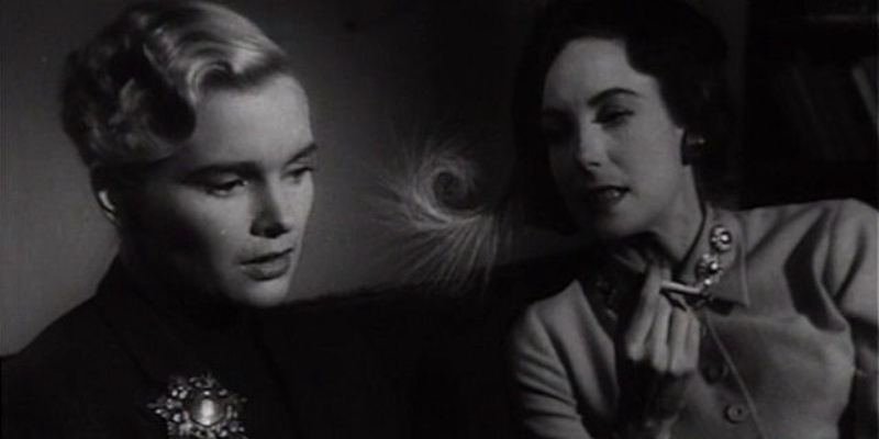 A blonde woman looks down and a brunette looks at her while smoking a cigarette. The image is Black and White and they're shrouded in shadows.