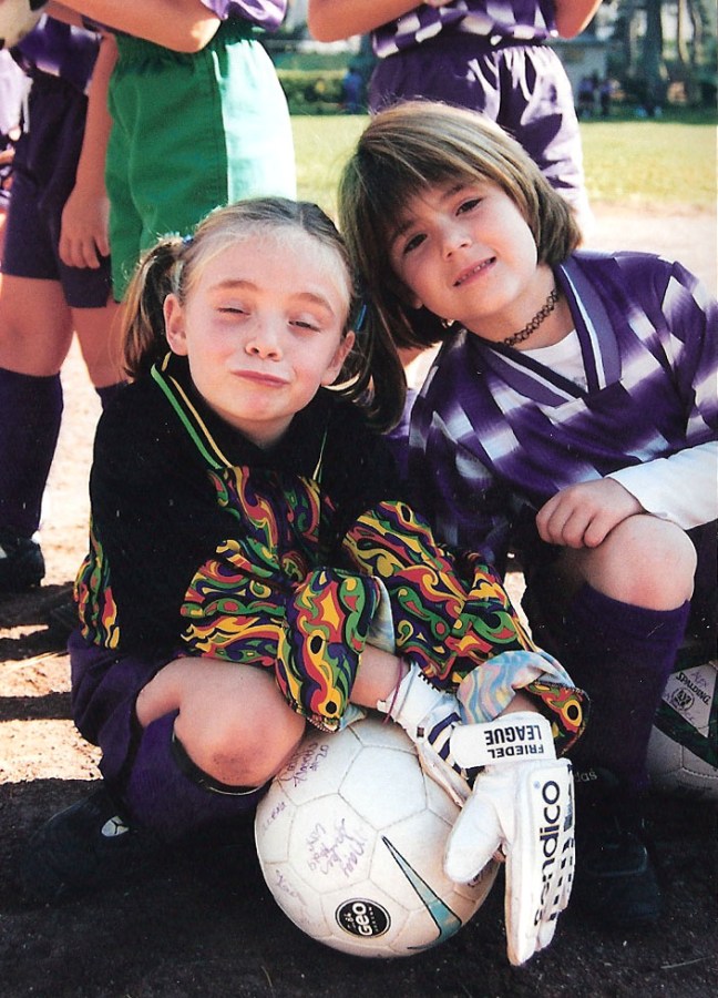 Two young girls kneel on the ground, dressed in football uniforms