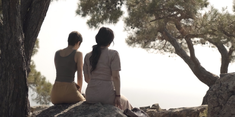 Two women sit on a rock with their backs to the camera looking out at a coast surrounded by trees.