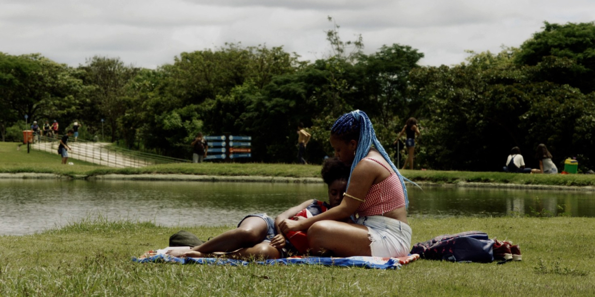 Mars One: Two young Black femmes cuddle on a blanket in a park.