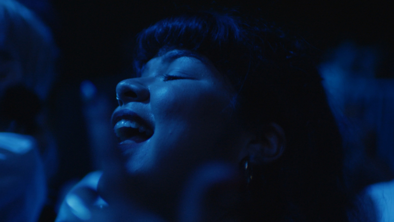 Close-up of Doris Muñoz's face in dark blue lighting. She is looking up with her mouth open as she dances.