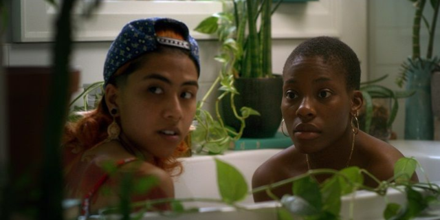 Noor and Layla sit in a bathtub surrounded by plants as they look towards something in the slight distance. queer muslim love