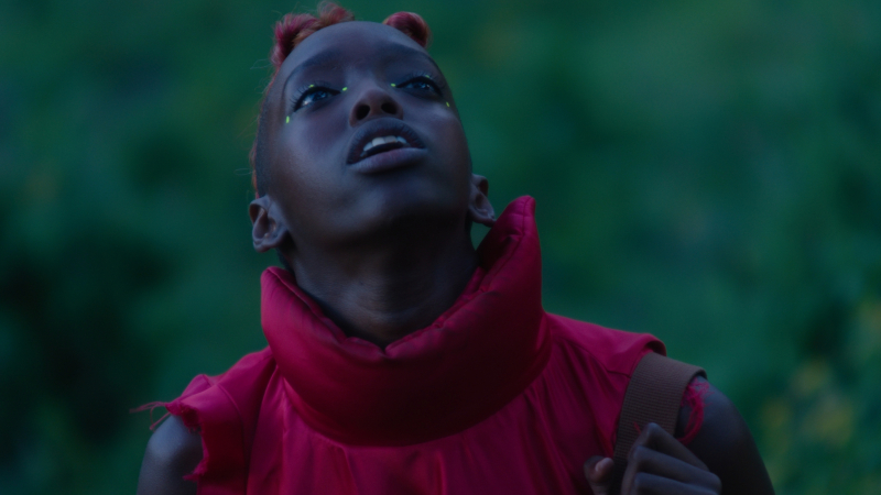 A woman wearing a puffy red vest with a collar looks up. Her eyes have neon green dots around them.