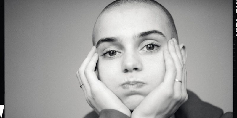 A black and white close up of young Sinead O'Connor as she puffs out her cheeks as she holds her face in her hands