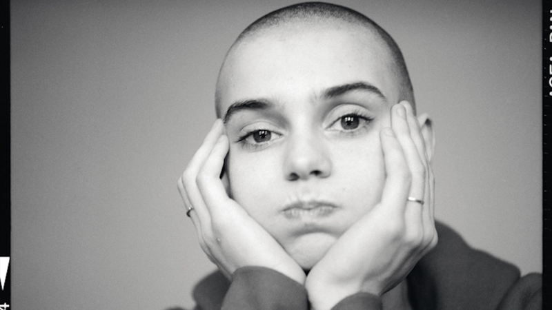A black and white close up of young Sinead O'Connor as she puffs out her cheeks as she holds her face in her hands