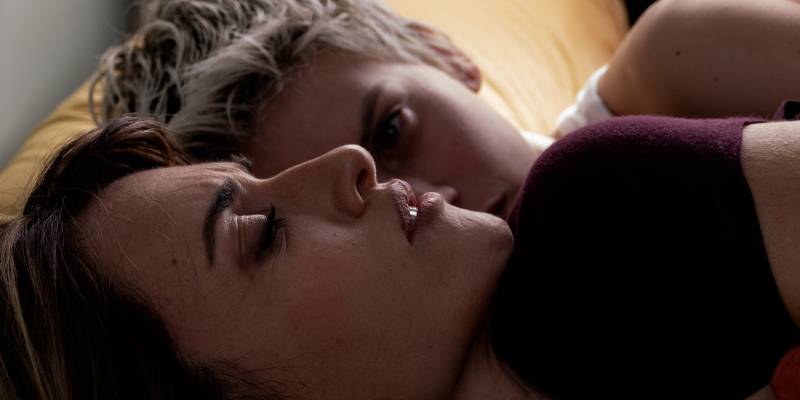 Close-up from the side of Penelope Cruz on her back with her eyes closed. A young woman with short blonde hair lies next to her watching.