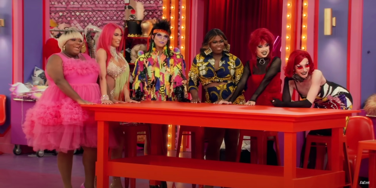 drag race recap 1403: Kornbread, Kerri, Alyssa, June, Willow, and Bosco stand around a table staring down the other queens with smiles.