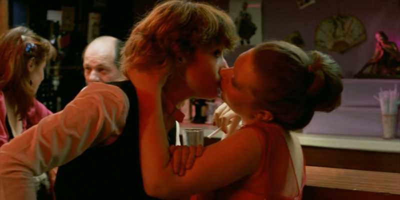 A woman in a dress pulls a woman in a dress shirt and black vest toward her for a kiss. A male bartender speaks to another woman in the background.